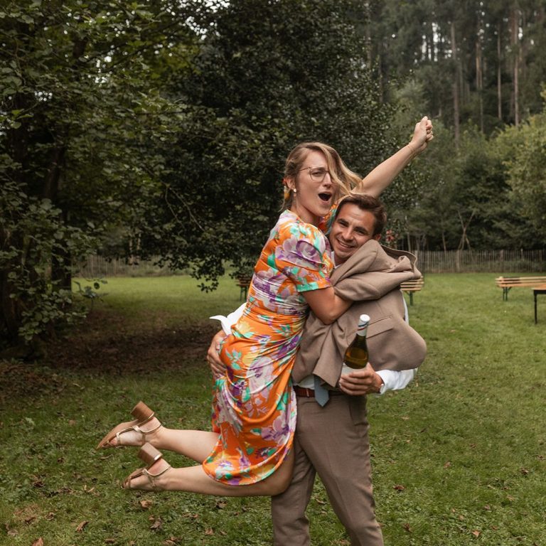 Une invitée de mariage souriante portée par un homme dans un champ entouré d'arbres.