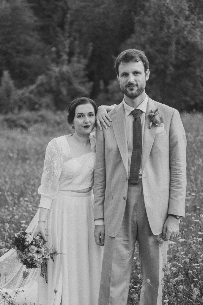 Jeunes mariés posant dans un champ fleuri, souriant, vêtus élégamment.