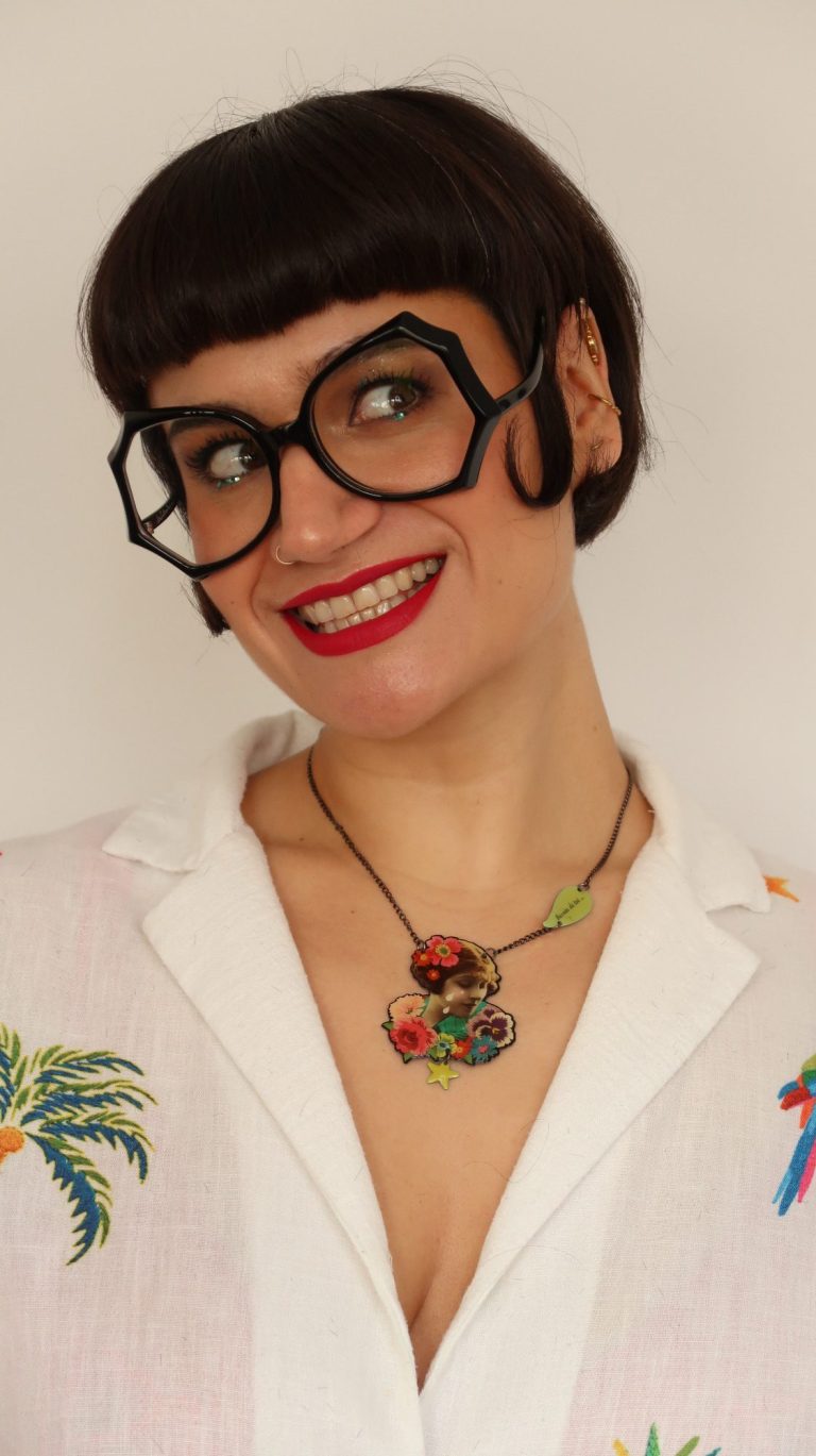 Femme souriante avec des lunettes noires épaisses et un collier coloré, portant une chemise blanche.